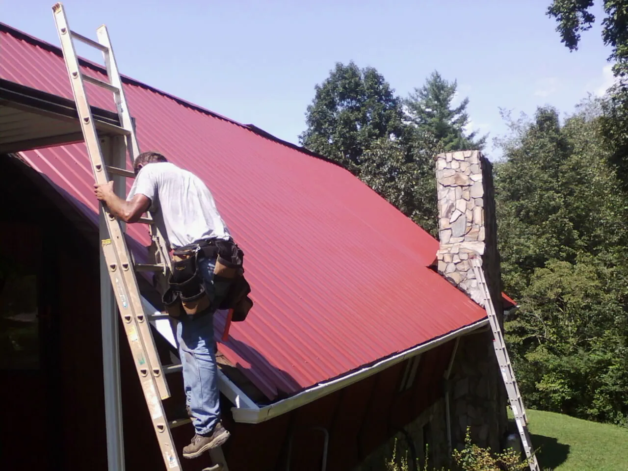 Roofer on a ladder beside a red metal roof and stone chimney — Soffit & Fascia Repair by Roofing Brothers in Asotin